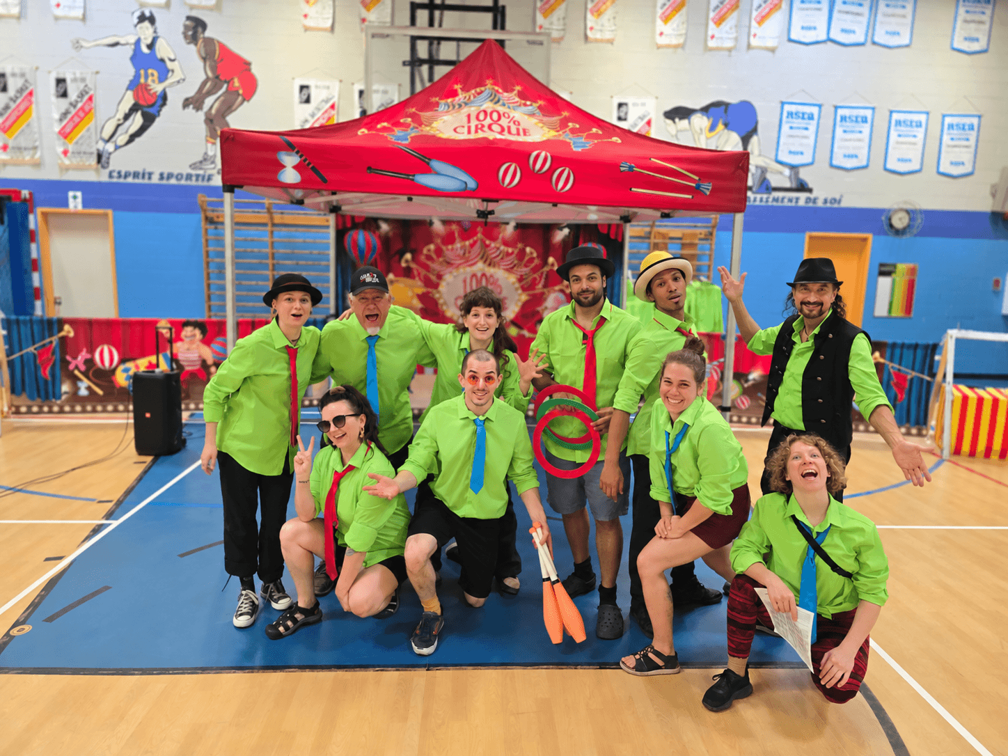 Toute l’équipe de 100 % Cirque souriante devant le chapiteau rouge et le décor de cirque dans un gymnase