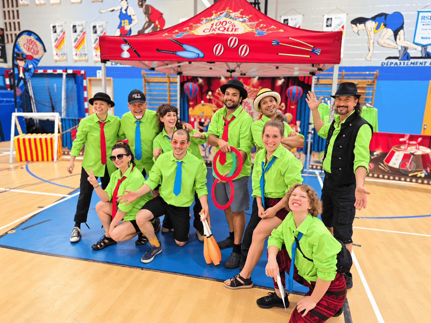 Toute l’équipe de 100 % Cirque souriante devant le chapiteau rouge et le décor de cirque dans un gymnase